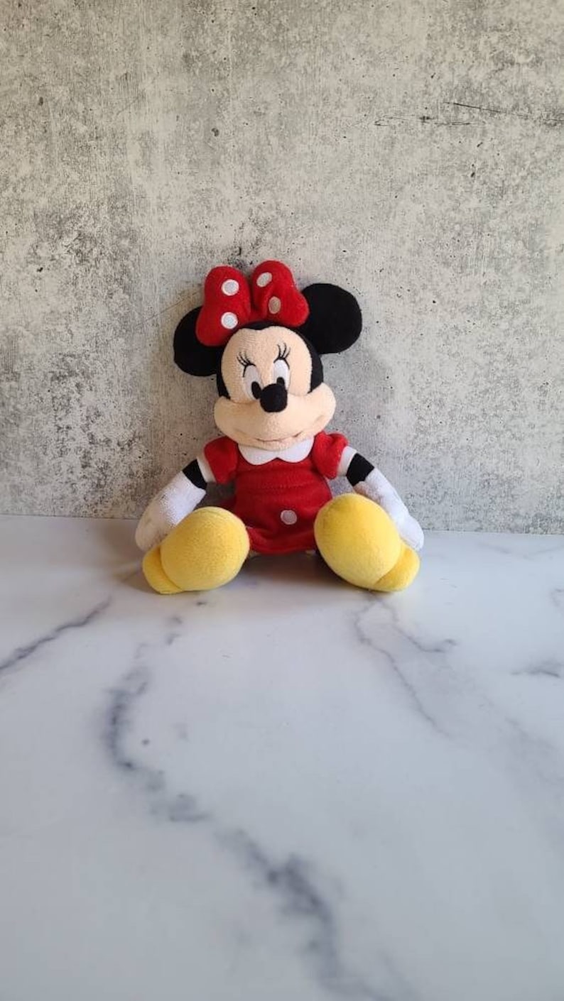 K&ouml;nnte beinhalten: Eine Pl&uuml;schpuppe von Minnie Mouse, mit schwarzen Ohren, einer roten Schleife mit wei&szlig;en Punkten und einem roten Kleid. Die Puppe hat gelbe Schuhe und wei&szlig;e Handschuhe und sitzt.