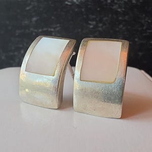 May include: A pair of silver-toned earrings with a rectangular, off-white mother-of-pearl inlay. The earrings have a curved, rectangular shape and a post backing. The background is a dark, blurred surface.