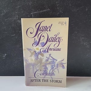 May include: A paperback book titled "Americana Colorado After The Storm" by Janet Dailey. The cover features a floral design in shades of purple and white against a beige background. The book is standing upright.