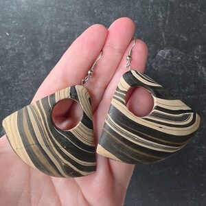 May include: A pair of dangle earrings featuring a curved, fan-like shape with a circular cutout. The earrings display a marbled pattern of black, tan, and cream swirls throughout the surface, attached to silver-toned metal earring hooks.