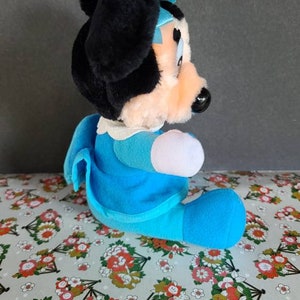 May include: A plush toy of a cartoon mouse wearing a blue dress and a blue bow. The toy is sitting on a floral patterned fabric.