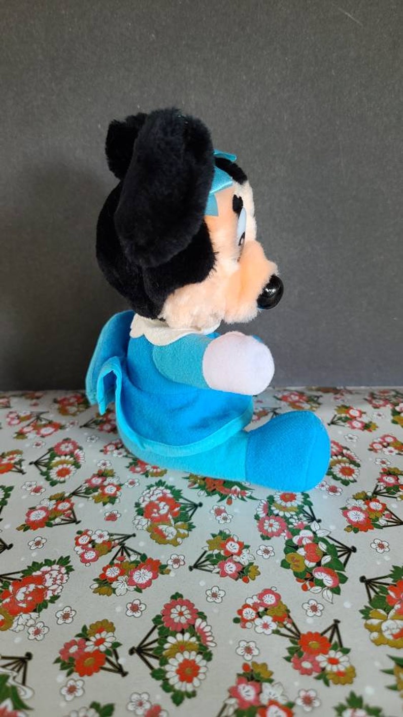 May include: A plush toy of a cartoon mouse wearing a blue dress and a blue bow. The toy is sitting on a floral patterned fabric.