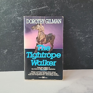 Puede incluir: Una portada de libro con un fondo morado que presenta un caballo de madera en una cuerda floja. El título del libro es "The Tightrope Walker" de Dorothy Gilman. El libro es de la autora de las famosas misterios de la Sra. Pollifax.