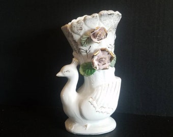 Puede incluir: Jarrón de cerámica blanca con forma de pavo real con rosas rosas y blancas en la parte superior. El jarrón tiene detalles dorados.