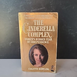 Puede incluir: Un libro de bolsillo dorado con el título "The Cinderella Complex: Women's Hidden Fear of Independence" de Colette Dowling. La portada presenta a una mujer con cabello castaño corto y una camisa blanca.