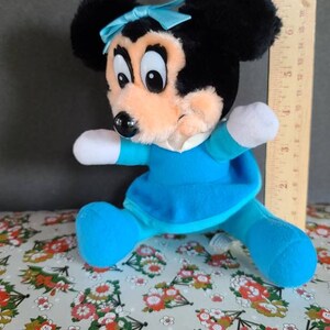 May include: A plush toy of a cartoon mouse wearing a blue dress and a blue bow. The mouse has black ears, a black nose, and large black eyes. The toy is about 7 inches tall.
