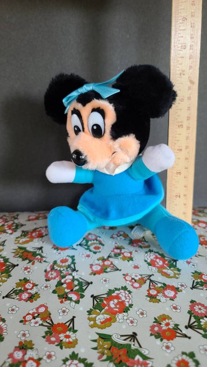 May include: A plush toy of a cartoon mouse wearing a blue dress and a blue bow. The mouse has black ears, a black nose, and large black eyes. The toy is about 7 inches tall.