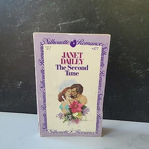 May include: A vintage paperback book titled "The Second Time" by Janet Dailey. The cover features an illustration of a couple embracing, with a floral arrangement. The book is part of the Silhouette Romance series.