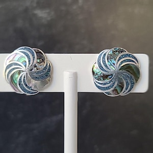 May include: A pair of silver-toned stud earrings with a floral design. Each earring features a central abalone shell surrounded by blue accents and silver-colored petals. The earrings are displayed on a white stand against a dark background.