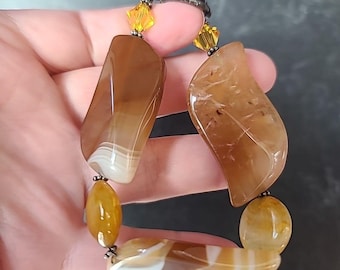 May include: A handcrafted bracelet featuring large, polished agate stones in shades of brown, cream, and amber. The bracelet includes smaller, rounded amber-colored beads and yellow crystal accents. The bracelet is held in a hand.
