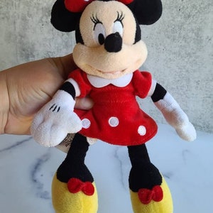 K&ouml;nnte beinhalten: Eine Pl&uuml;schpuppe von Minnie Mouse. Sie tr&auml;gt ein rotes Kleid mit wei&szlig;en Punkten, einen wei&szlig;en Kragen und eine rote Schleife mit wei&szlig;en Punkten. Ihre Schuhe sind gelb mit roten Schleifen. Die Puppe hat schwarze Ohren und eine schwarze Nase.