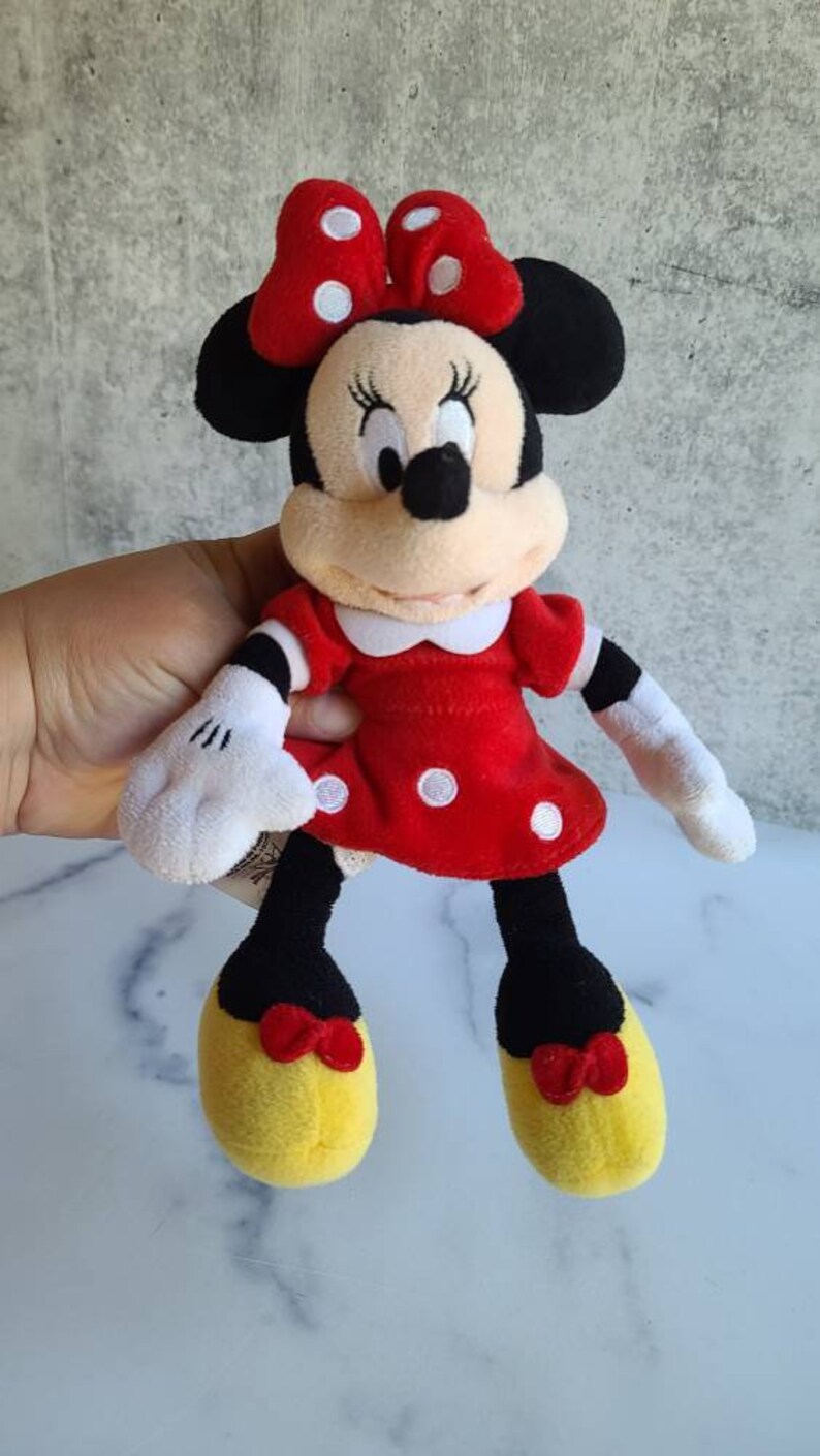 K&ouml;nnte beinhalten: Eine Pl&uuml;schpuppe von Minnie Mouse. Sie tr&auml;gt ein rotes Kleid mit wei&szlig;en Punkten, einen wei&szlig;en Kragen und eine rote Schleife mit wei&szlig;en Punkten. Ihre Schuhe sind gelb mit roten Schleifen. Die Puppe hat schwarze Ohren und eine schwarze Nase.