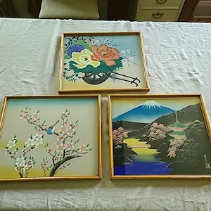 May include: Three framed prints with gold frames. The first print features a floral arrangement with a cart and wheels. The second print features a bird in a tree with white blossoms. The third print features a mountain scene with a river and trees.