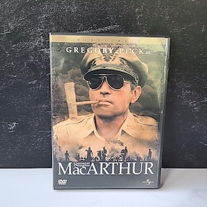 以下が含まれることがあります： グレゴリー・ペック主演の映画「マッカーサー」のDVD。カバーには、軍服とサングラスを着用したペックの肖像画が描かれており、その下にタイトルが大きく表示されています。DVDケースはビニールスリーブに入っています。