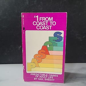 May include: A vintage paperback book with a magenta cover. The title "#1 FROM COAST TO COAST" is displayed in white, with the word "PASSAGES" in colorful stacked letters. The book's subtitle reads "PREDICTABLE CRISES OF ADULT LIFE" by Gail Sheehy.