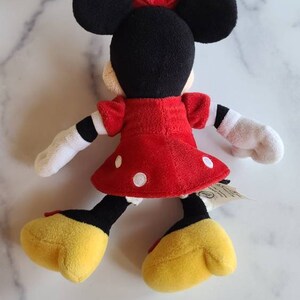 K&ouml;nnte beinhalten: Pl&uuml;schpuppe von Minnie Mouse, von hinten gesehen, mit einem roten Kleid mit wei&szlig;en Punkten, schwarzen Ohren und einer roten Schleife. Die Puppe hat wei&szlig;e Handschuhe, gelbe Schuhe und schwarze und gelbe Beine. Ein klassischer Disney-Charakter.