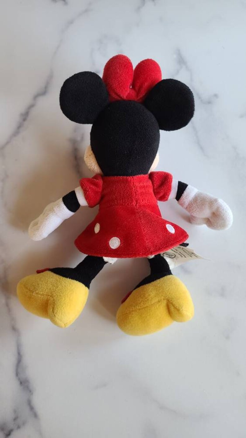 K&ouml;nnte beinhalten: Pl&uuml;schpuppe von Minnie Mouse, von hinten gesehen, mit einem roten Kleid mit wei&szlig;en Punkten, schwarzen Ohren und einer roten Schleife. Die Puppe hat wei&szlig;e Handschuhe, gelbe Schuhe und schwarze und gelbe Beine. Ein klassischer Disney-Charakter.