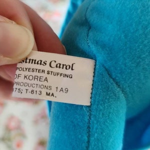 May include: A white tag with the text "stmas Carol POLYESTER STUFFING OF KOREA PRODUCTIONS 1A9 175; T-613 MA." attached to a blue fabric item.