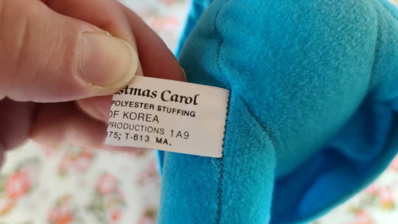 May include: A white tag with the text "stmas Carol POLYESTER STUFFING OF KOREA PRODUCTIONS 1A9 175; T-613 MA." attached to a blue fabric item.