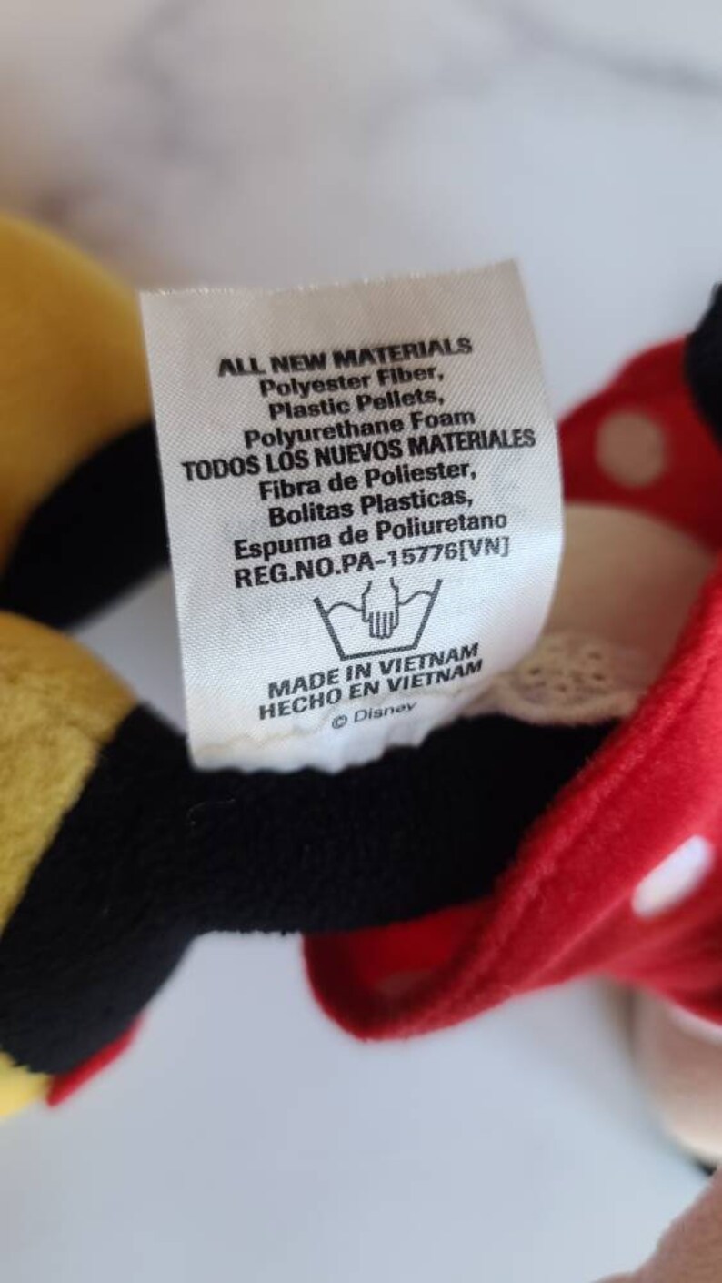 K&ouml;nnte beinhalten: Ein Etikett von einem Disney-Pl&uuml;schtier mit Text in Englisch und Spanisch. Das Etikett listet Materialien wie Polyesterfaser und Polyurethanschaum auf. Das Etikett gibt auch an, dass das Spielzeug in Vietnam hergestellt wurde.