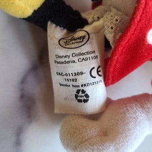 K&ouml;nnte beinhalten: Nahaufnahme eines Disney Collection Pl&uuml;schtier-Etiketts. Das Etikett enth&auml;lt das Disney Collection-Logo, die Adresse und Artikeldetails. Das Pl&uuml;schtier hat eine rote Schleife mit wei&szlig;en Punkten, gelbe und schwarze Details und eine wei&szlig;e Pl&uuml;schhand.