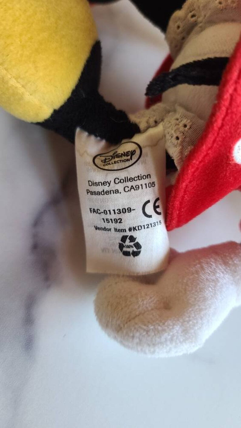 K&ouml;nnte beinhalten: Nahaufnahme eines Disney Collection Pl&uuml;schtier-Etiketts. Das Etikett enth&auml;lt das Disney Collection-Logo, die Adresse und Artikeldetails. Das Pl&uuml;schtier hat eine rote Schleife mit wei&szlig;en Punkten, gelbe und schwarze Details und eine wei&szlig;e Pl&uuml;schhand.