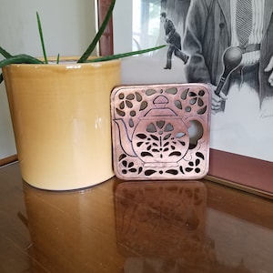 Puede incluir: Un posavasos de metal de color cobre en forma de tetera con un diseño recortado. El posavasos está sobre una superficie de madera.