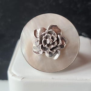 May include: A silver-colored ring with a flower design. The flower is centered on a round, off-white, mother-of-pearl-like surface. The flower has detailed petals and a textured appearance. The ring is displayed on a white surface.