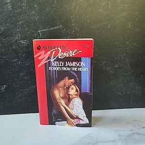 Puede incluir: Un libro de bolsillo titulado "Echoes From The Heart" de Kelly Jamison. La portada presenta una pareja abrazándose, con el hombre sin camisa. El libro forma parte de la serie Silhouette Desire, con una combinación de colores rojo y blanco.