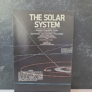 Puede incluir: Una portada vintage de la revista National Geographic titulada "The Solar System". La portada presenta un fondo oscuro con líneas y texto blancos, incluyendo "Jupiter" y datos científicos. Publicado en julio de 1981.