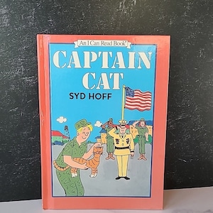 Puede incluir: Un libro infantil vintage titulado "Captain Cat" de Syd Hoff. La portada presenta una ilustración de dibujos animados de un gato con uniforme militar sosteniendo un gatito. El libro forma parte de la serie "An I Can Read Book".
