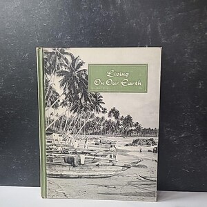 Puede incluir: Un libro de tapa dura titulado "Living On Our Earth" con lomo verde. La portada presenta una ilustración en blanco y negro de una escena de playa con palmeras, botes y personas. El libro está sobre un fondo oscuro.