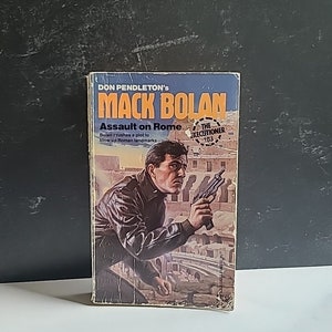 May include: A paperback book cover featuring a man holding a gun in front of a Roman Colosseum. The book title is "Mack Bolan: Assault on Rome" and the subtitle is "Bolan crushes a plot to blow up Roman landmarks". The book is part of "The Executioner" series, number 103.