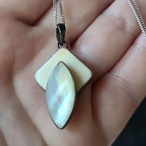 以下が含まれることがあります： クリーム色の正方形と、洋梨型の虹色の真珠母貝の石を特徴とするペンダントが付いたシルバーのネックレス。ペンダントは繊細なシルバーチェーンから吊り下げられています。ネックレスはニュートラルな背景に表示されています。