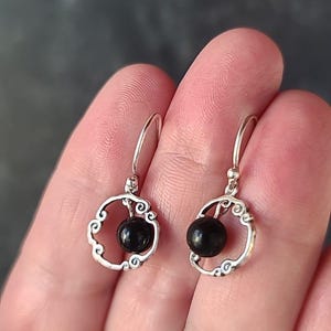 Puede incluir: Un par de pendientes colgantes de plata. Cada pendiente presenta una pequeña cuenta negra redonda engastada en un marco decorativo calado de plata. El marco tiene un diseño ornamental y arremolinado. Los pendientes se muestran sobre un fondo neutro.