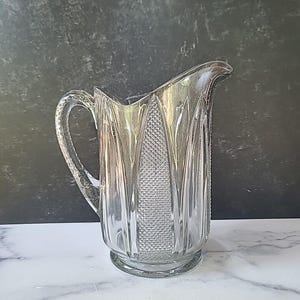 May include: A clear glass pitcher with a textured design and a handle. The pitcher has a wide mouth and a round base.