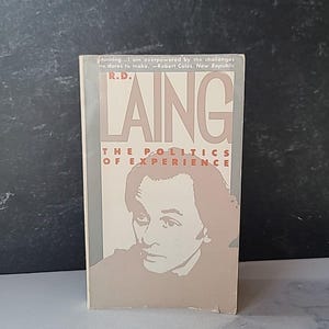 May include: A paperback book titled "R.D. Laing The Politics of Experience". The cover features a portrait of a man in a light brown color, with the author's name in large, bold letters. The book is set against a dark background.
