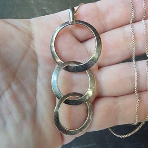 May include: A silver-toned necklace featuring a pendant composed of three interlocking circular rings of varying sizes, suspended from a delicate box chain. The metal surface displays a subtle, aged patina with visible signs of wear.