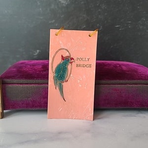 May include: A pink tag with a gold border and a blue and green parrot illustration. The tag reads "Polly Bridge".