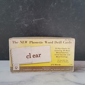以下が含まれることがあります： 「The NEW Phonetic Word Drill Cards」と題された、ヴィンテージの教育用カードセット。箱はクリーム色で、茶色の文字とグラフィックが施されています。「clear」という単語が表示されたカードが1枚あります。このセットには、単語ファミリーを学ぶための10個のフリップチャートが含まれています。