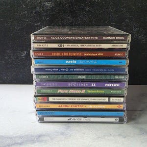 以下が含まれることがあります： 透明なプラスチックケースに入った12枚の音楽CDの垂直スタック。背表紙には、アリス・クーパー、キッス、フーティー＆ザ・ブロウフィッシュ、オアシス、ミッシー・エリオット、グリース、ボーイズIIメン、ピュア・ディスコ2、クランベリーズ、アーロン・カーター、ウィーザー、アヴィーチーなどのアーティスト名やアルバムタイトルが表示されています。