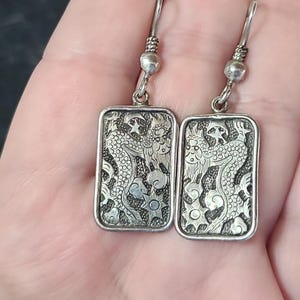 May include: A pair of silver-toned rectangular dangle earrings. Each earring features an engraved dragon design with clouds and stars. The earrings have a hook closure and are displayed against a neutral background.