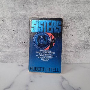 May include: A blue paperback book titled "Sisters" by Robert Littell. The cover features a man holding a rifle and the Central Intelligence Agency logo.