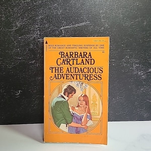 Puede incluir: Un libro de bolsillo amarillo con una pareja en la portada. El libro se titula "The Audacious Adventuress" de Barbara Cartland.