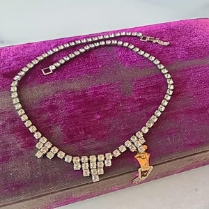 Puede incluir: Un collar de pedrería plateada con un diseño puntiagudo en forma de V en el centro. El collar se muestra sobre una superficie de terciopelo morado.