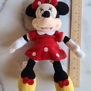 K&ouml;nnte beinhalten: Minnie Mouse Pl&uuml;schpuppe, mit einem roten Kleid mit wei&szlig;en Punkten, einer passenden Schleife und gelben Schuhen. Die Puppe hat schwarze Ohren und einen wei&szlig;en Kragen. Ein Holzlineal liegt neben der Puppe.