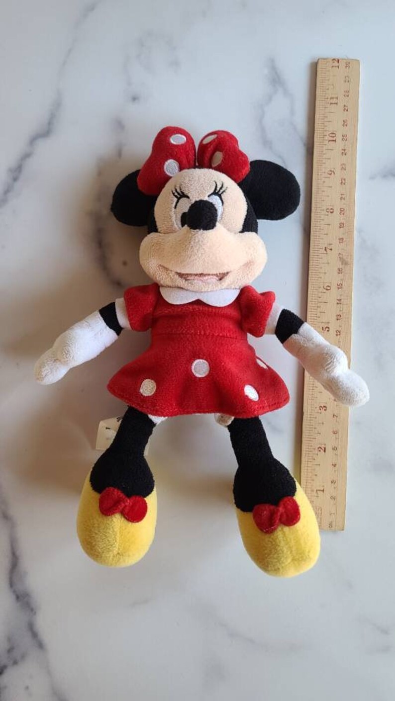 K&ouml;nnte beinhalten: Minnie Mouse Pl&uuml;schpuppe, mit einem roten Kleid mit wei&szlig;en Punkten, einer passenden Schleife und gelben Schuhen. Die Puppe hat schwarze Ohren und einen wei&szlig;en Kragen. Ein Holzlineal liegt neben der Puppe.