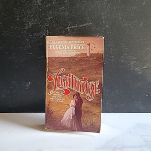 May include: A paperback book cover with a lighthouse and a couple embracing on the beach. The title of the book is "The Lighthouse" by Eugenia Price, the bestselling author of "The Beloved Invader" and "New Moon Rising".