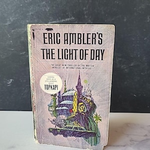 May include: A vintage paperback book cover featuring a colorful illustration of a cityscape with a car and a man in a cloak. The title of the book is "Eric Ambler's The Light of Day" and the text "The Great New Thriller by the Master Novelist of International Intrigue!" is printed below the title. The text "Coming Soon as a Spellbinding Movie Topkapi" is printed in a box at the bottom of the cover.