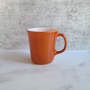 Puede incluir: Una taza naranja vintage con interior blanco. La taza tiene una forma redondeada y un asa.